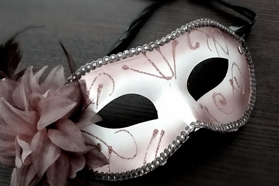 White and pink masquerade mask with feathers and a flower lying on a dark wooden surface, a symbol of bluffing in poker